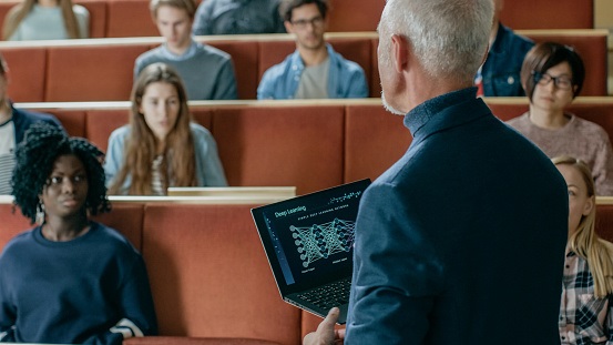Professor of Computer Science Reads Lecture to a Classroom Full of Multi Ethnic Students. Teacher Holds Laptop with Deep Learning, Artificial Intelligence Infographics on the Screen. Companies Bringing AI Training Inside; Udacity Well-Positioned 1